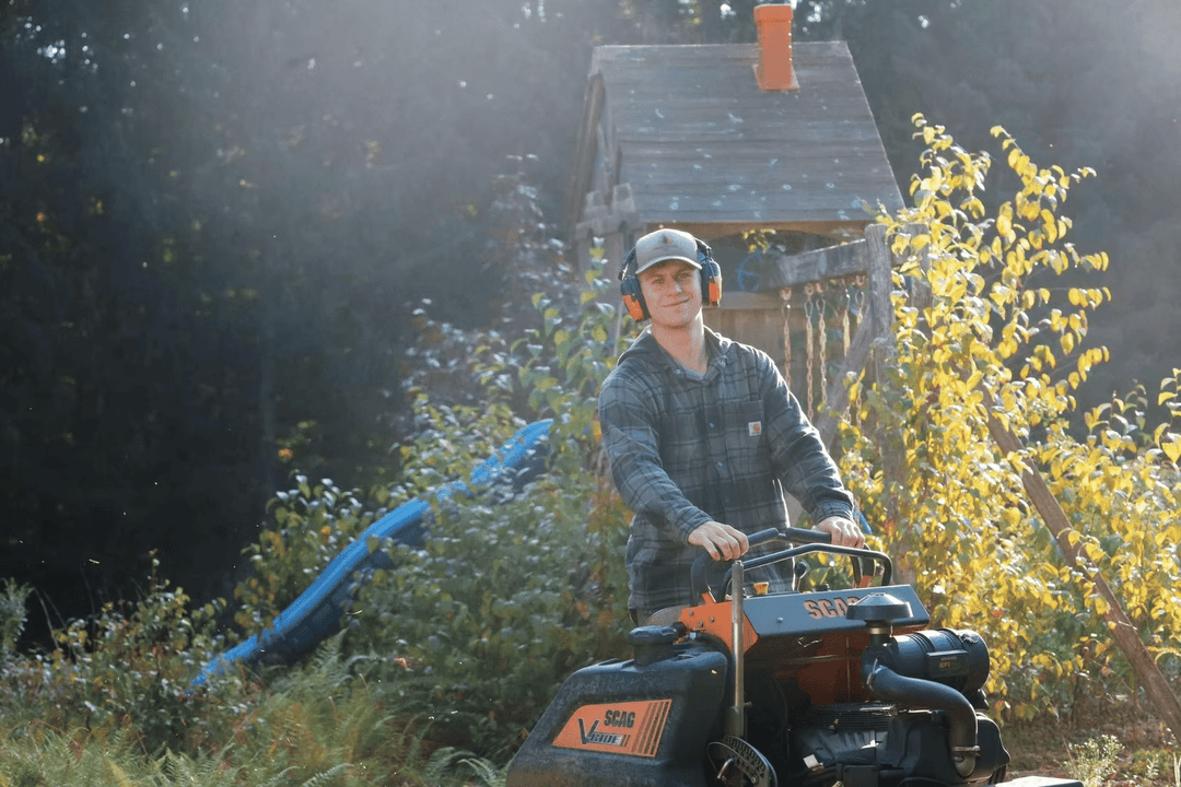 Man operating a lawn mower near a children’s playhouse in a sunlit garden.