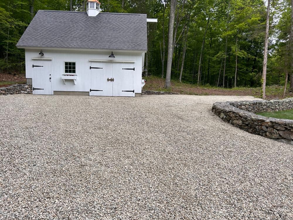 White barn-style garage surrounded by gravel driveway and stone landscaping in wooded area.