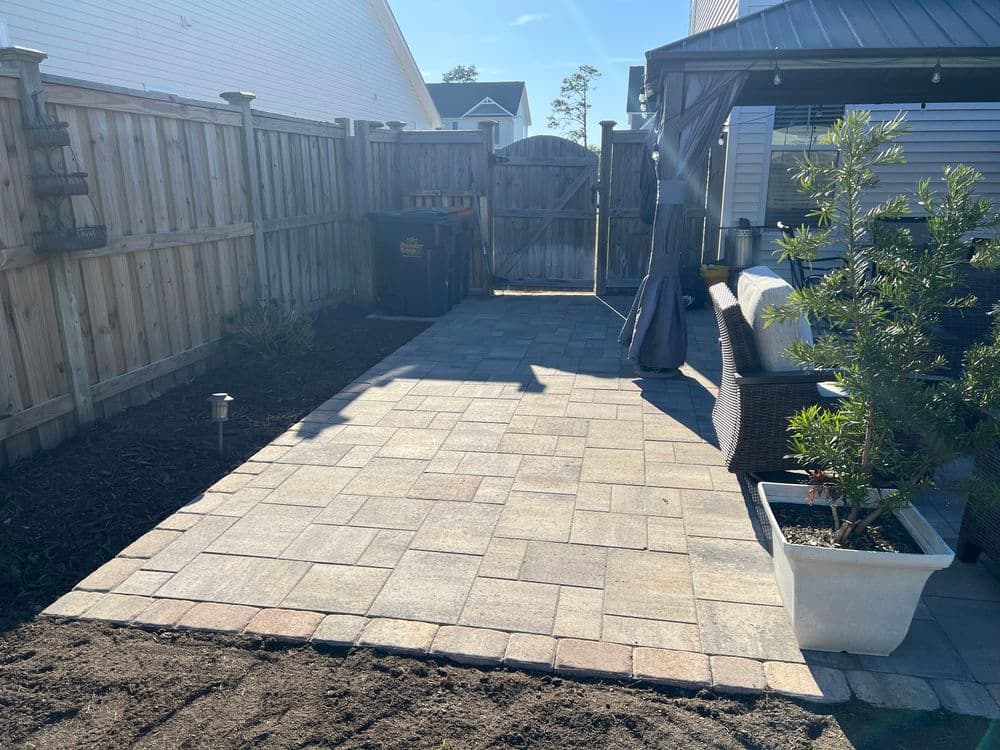 Paved backyard patio area with landscaping, seating, and a garden shed in sunlight.