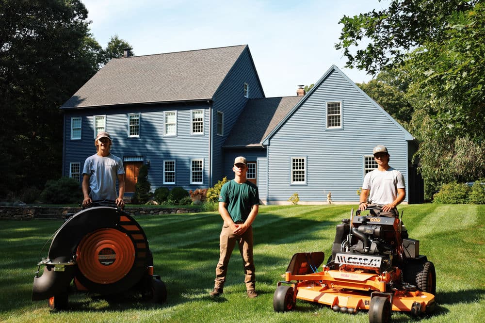 Landscaping professionals with equipment in front of a blue house on a lush green lawn.