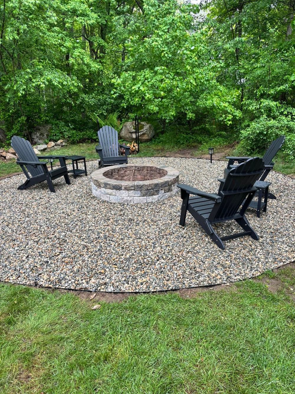 Outdoor fire pit surrounded by black adirondack chairs in lush green forest setting.