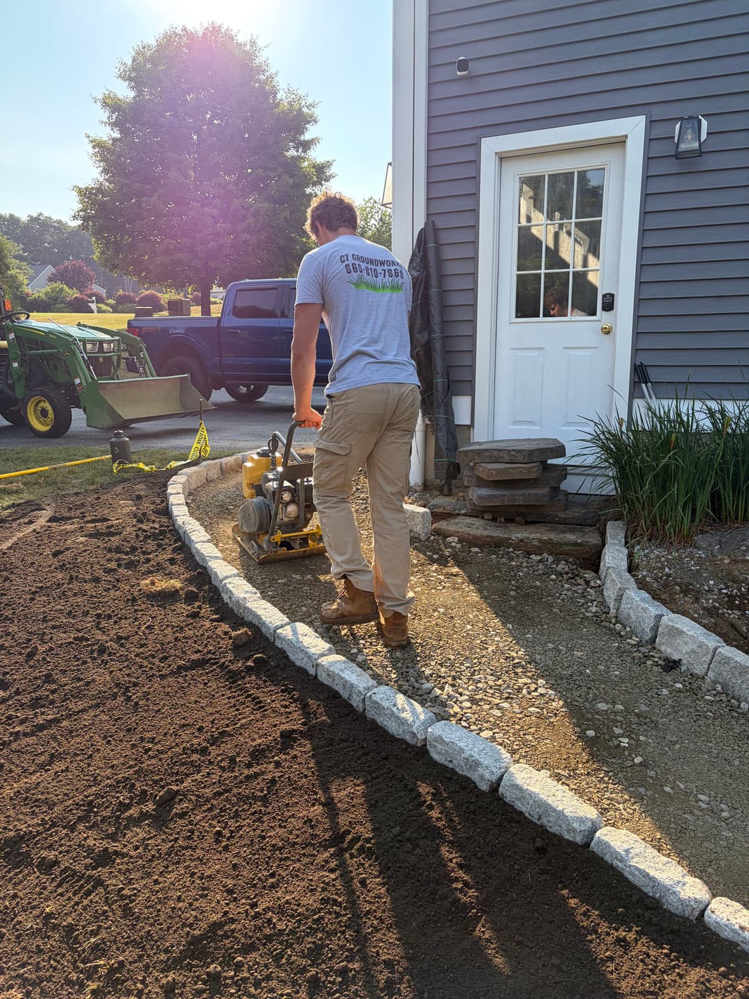 Man operating a plate compactor on a gravel walkway beside a house.