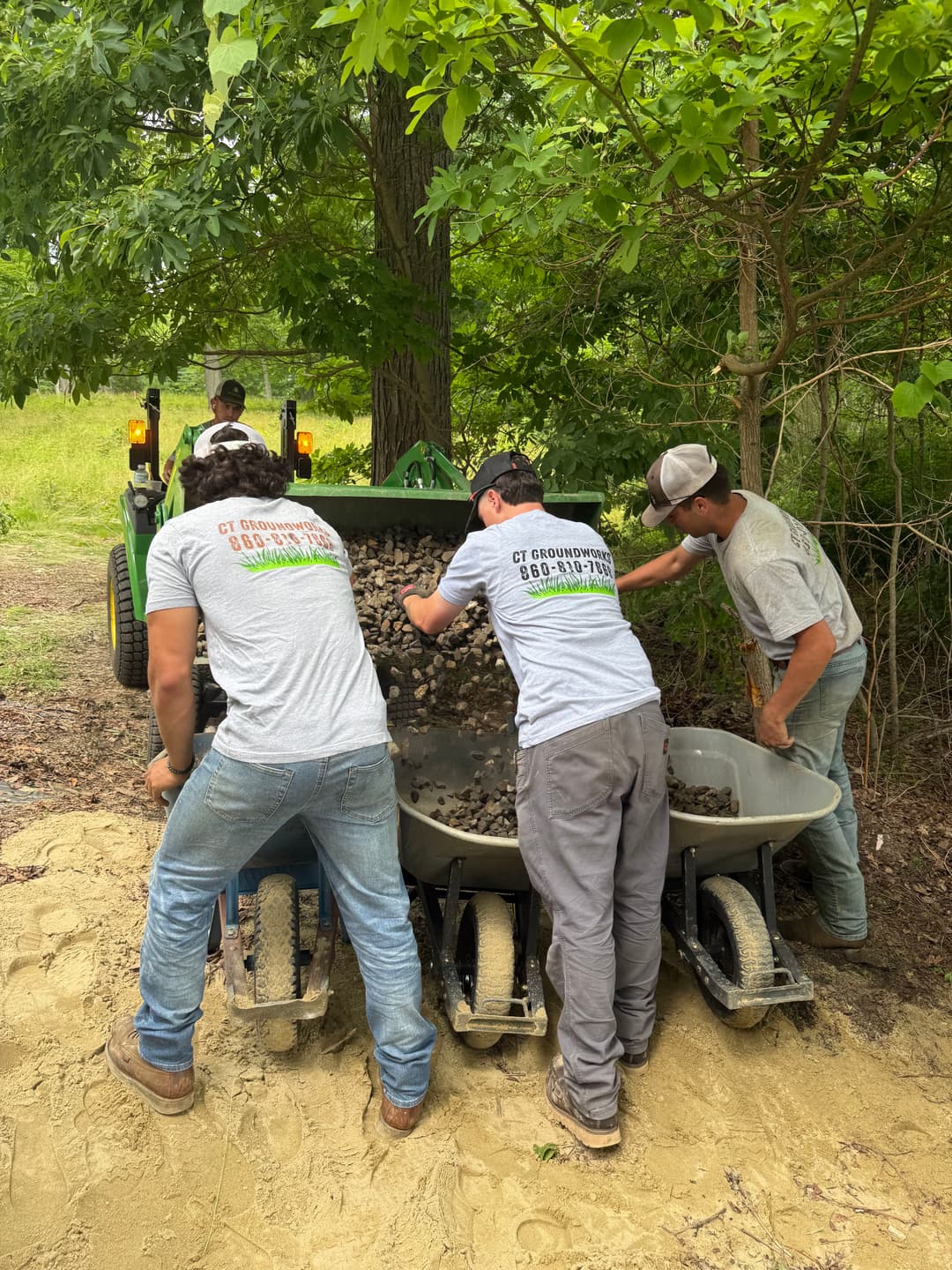 Workers loading wheelbarrows with gravel while operating machinery in a lush, green environment.