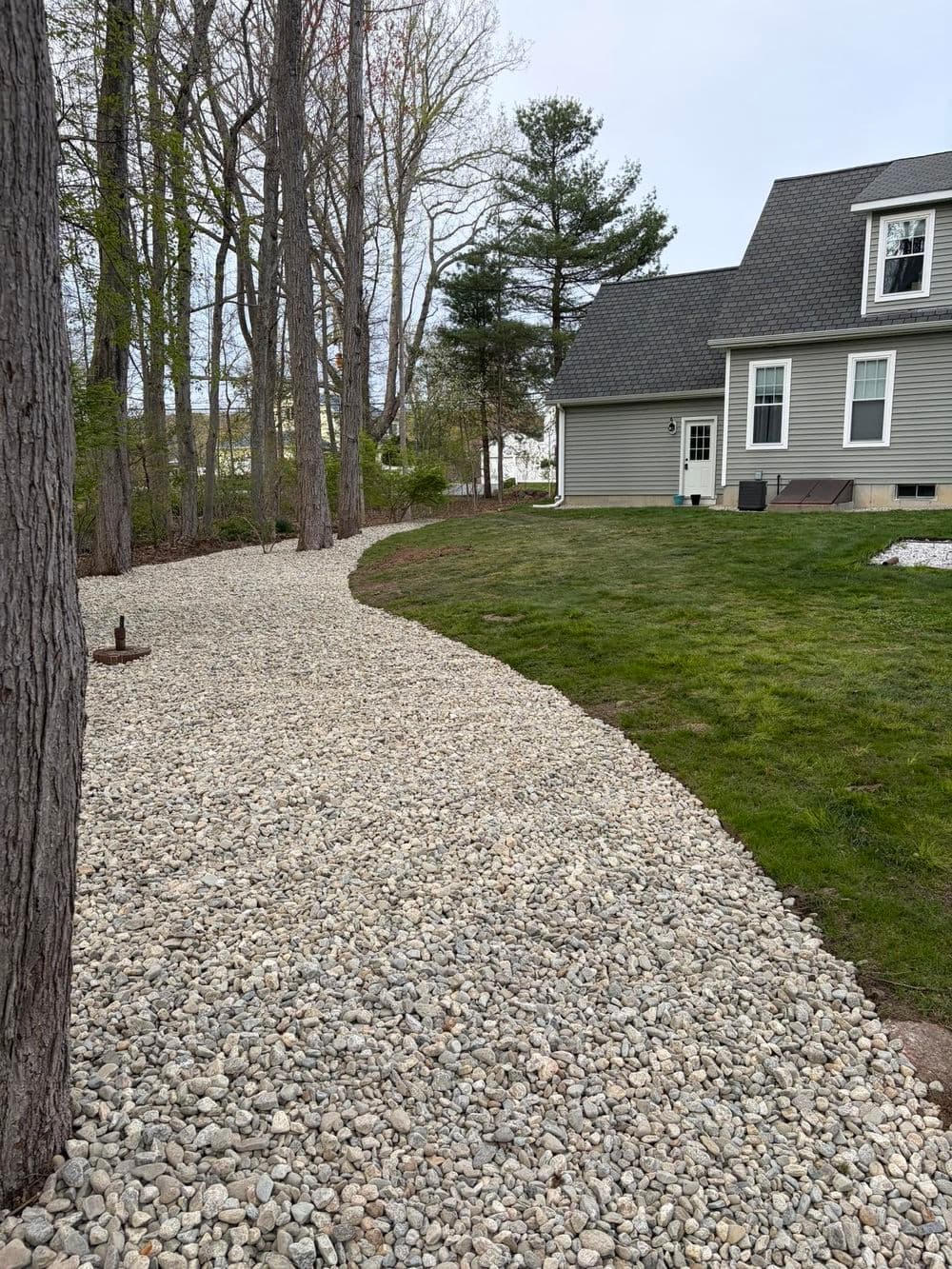 Gravel pathway leading to a gray house surrounded by trees and green grass.