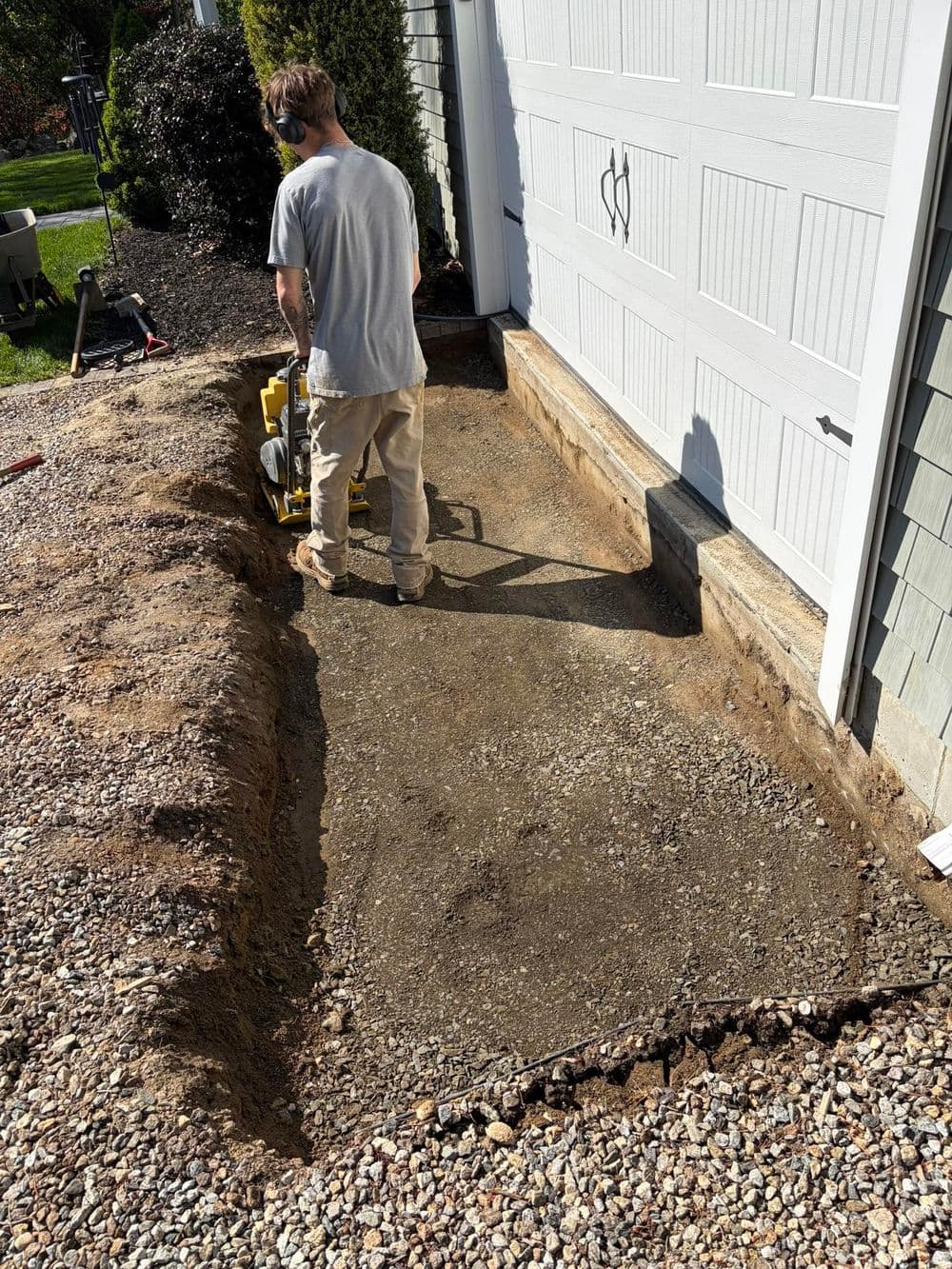 Person operating a plate compactor on a gravel surface near a garage.